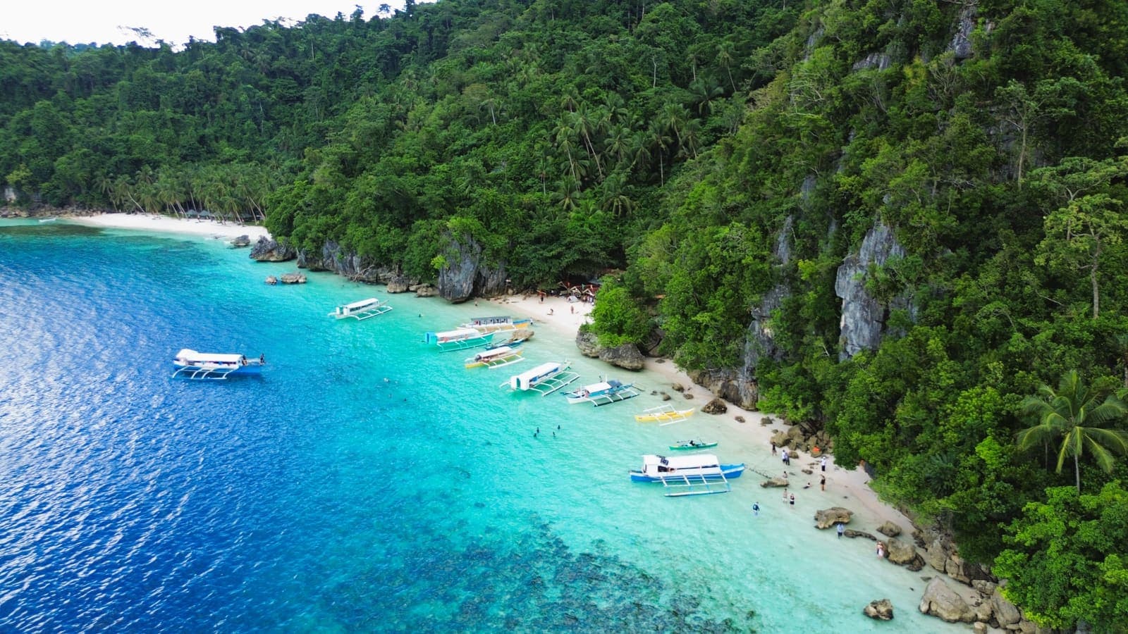 Outrigger boats moored off a white sand beach at Bitaog, backed by limestone cliffs and dense jungle