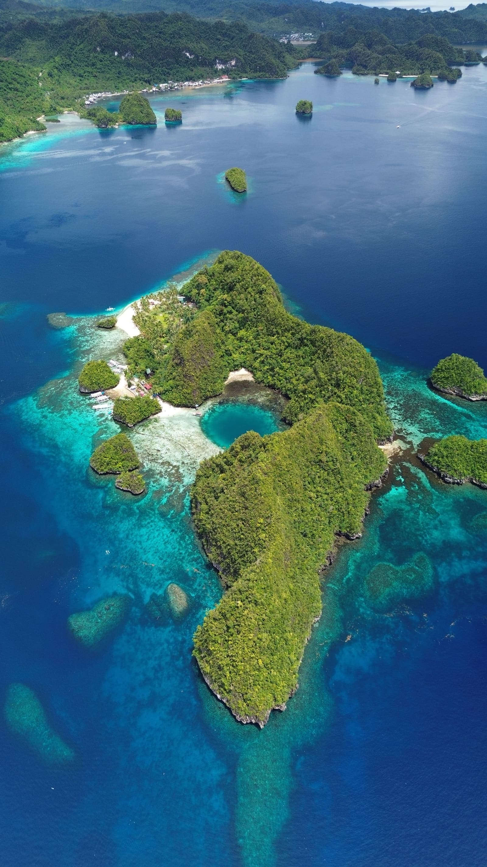 Aerial view looking down the length of a forested island in Dinagat, ringed by turquoise reef and deep blue channels