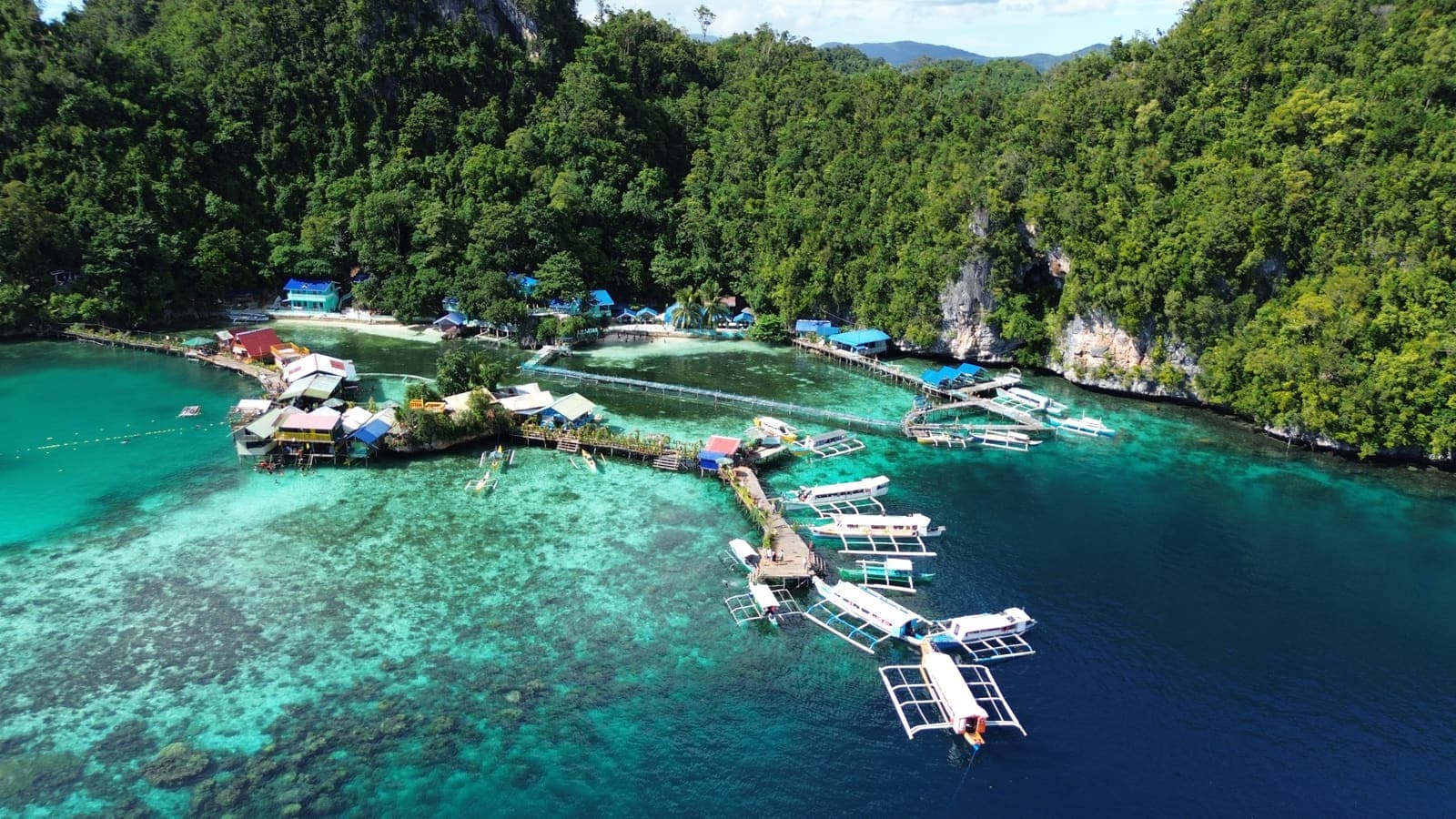 Outrigger boats moored in a row along the shore of a small island community, with clear reef visible beneath them