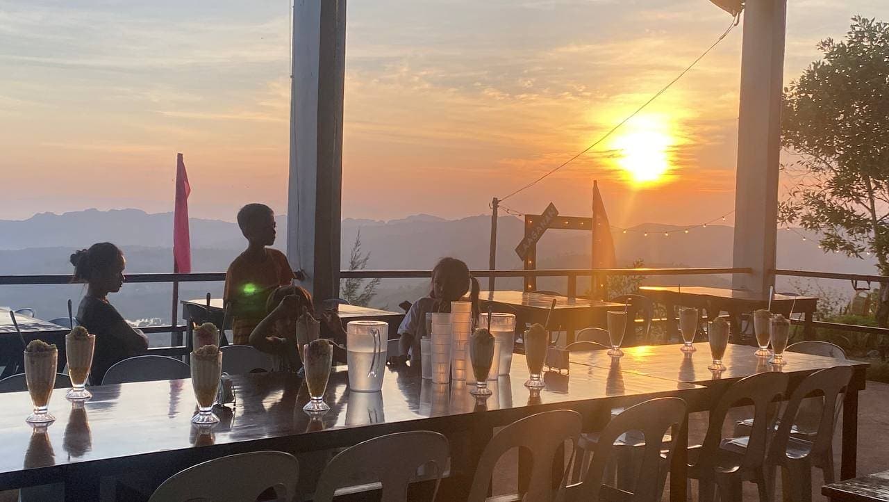 Guests seated at a long table on an open deck as the sun sets over distant mountains and sea